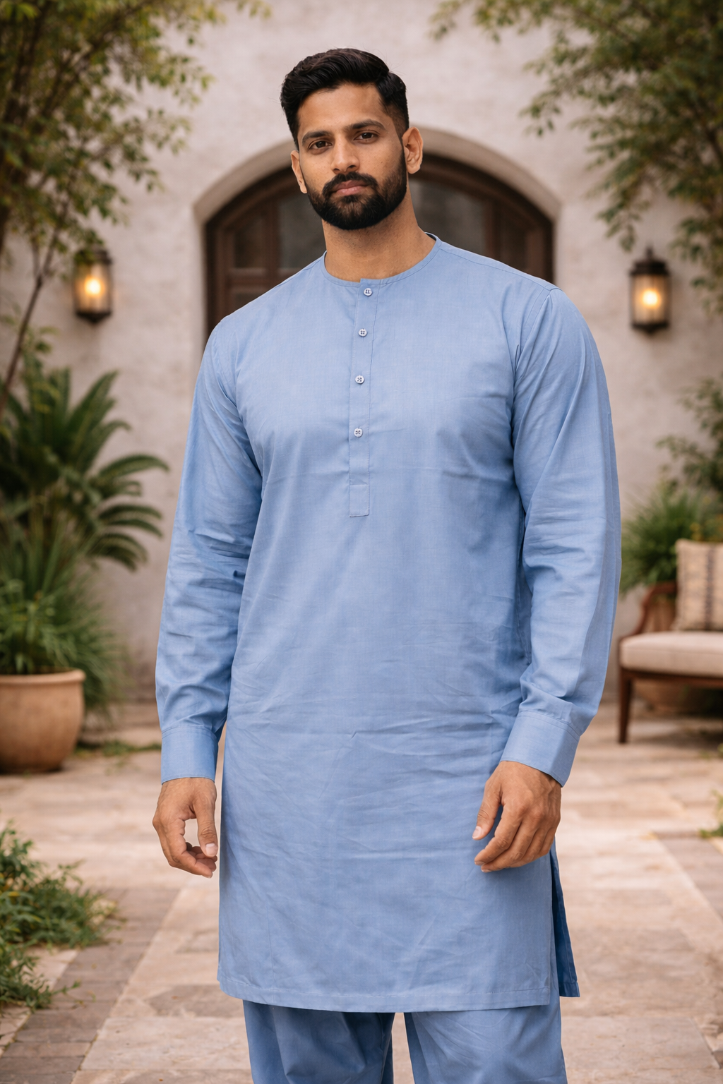 Man wearing a light blue shalwar kameez standing outdoors with plants and a building in the background